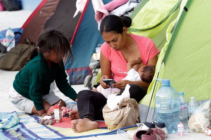 981428_Acajete Migrantes-6_web Mireya Novo Cuartoscuro A migrant mother sitting in the entrance to a family sized green camping tent holds a nursing baby in one arm while she checks her cell phone. Her five-year-old daughter crouches in front of her. (Mireya Novo/Cuartoscuro)