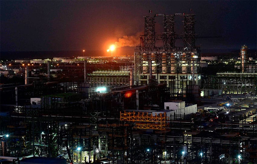 A gas flare burns at night in the middle of a large industrial complex, with the dark sea barely visible in the background.