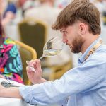Concours Mondial de Bruxelles wine taster seated in a large hall in Leon, Mexico, samples a white wine from a wine glass.