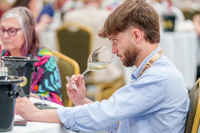 Taster from France Concours Mondial Concours Mondial de Bruxelles wine taster seated in a large hall in Leon, Mexico, samples a white wine from a wine glass.