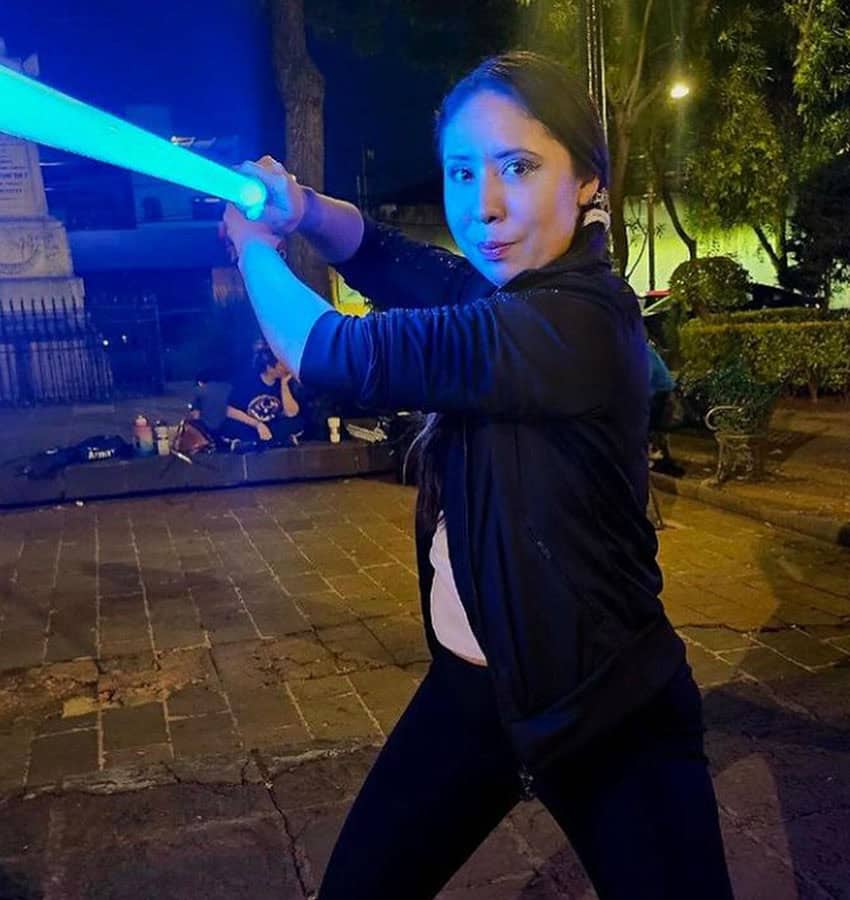 Woman posing in a fencing stance with a blue lightsaber in a public courtyard in Mexico City at night.